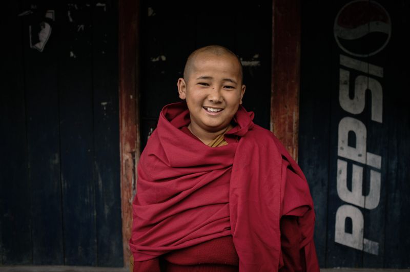 A young novice monk wrapped in a red robe, smiling, in front of a dark wooden door painted with a faded Pepsi logo.